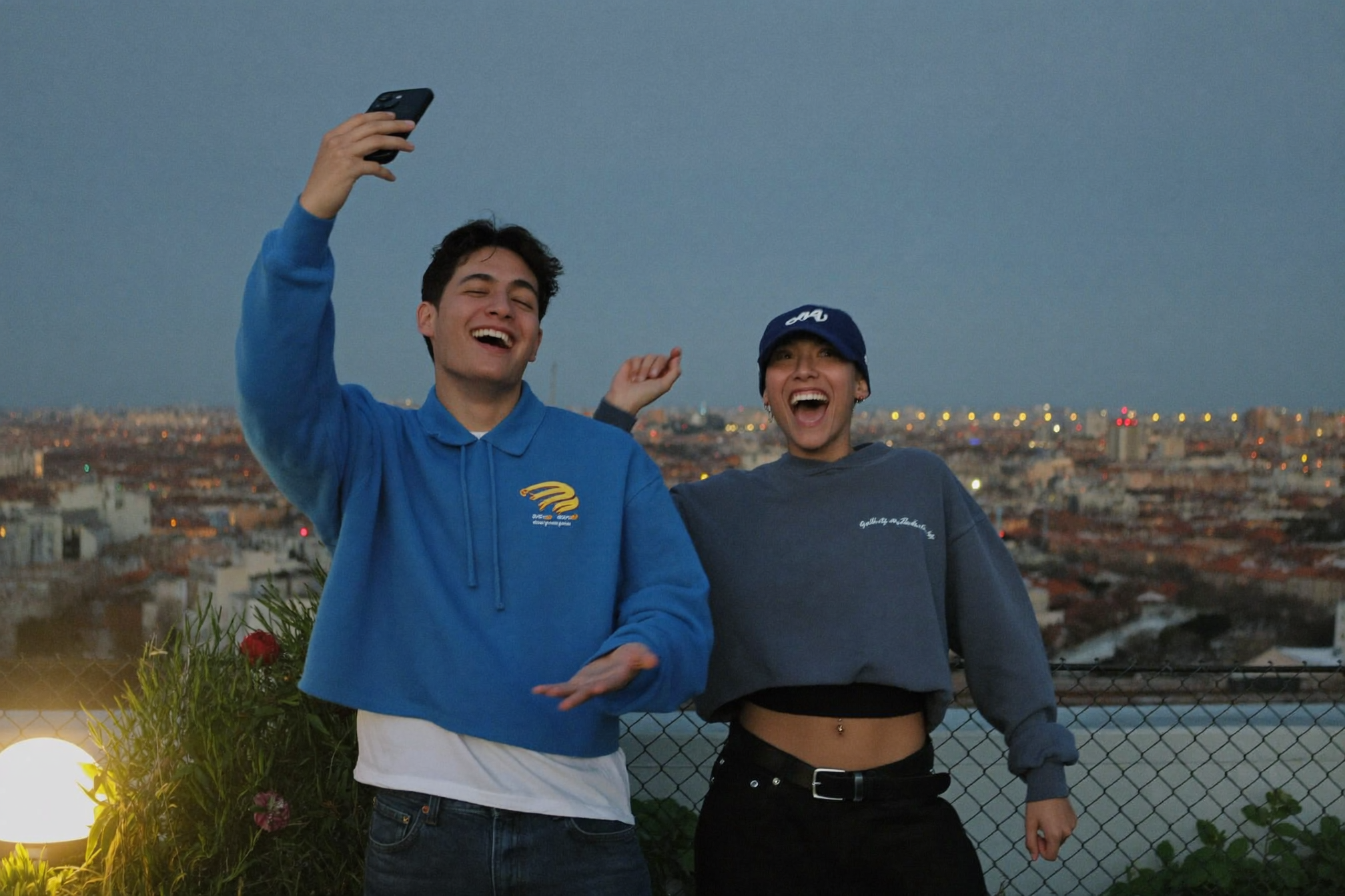 Two friends filming a trend on a rooftop at dusk, with the city lighting up behind them